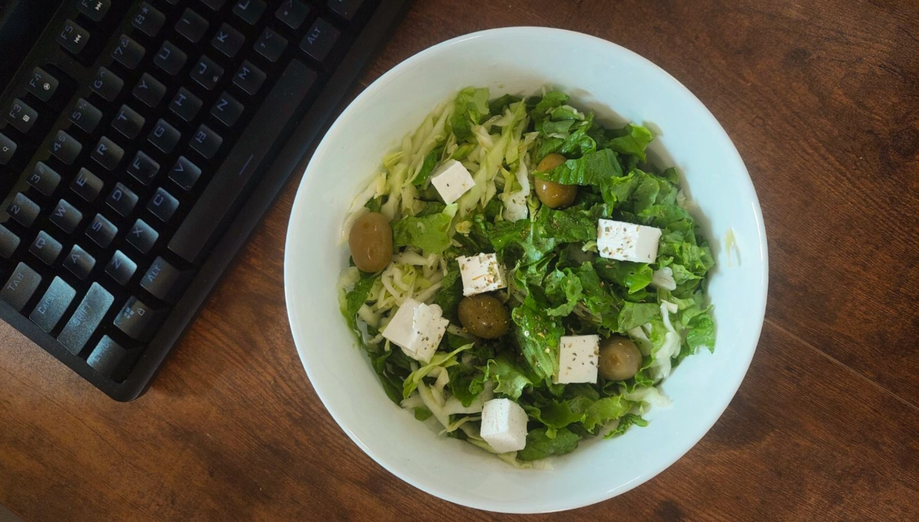 Mixed lettuce and cabbage salad in a bowl. Placed on a brown table, there is a keybord nex to it.