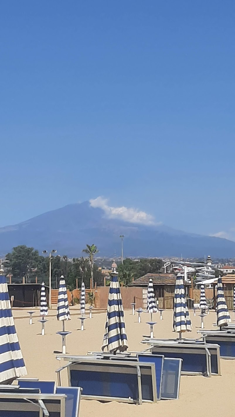 A beach with blue-and-white striped umbrellas and Mount Etna smoking in the distance in Catania