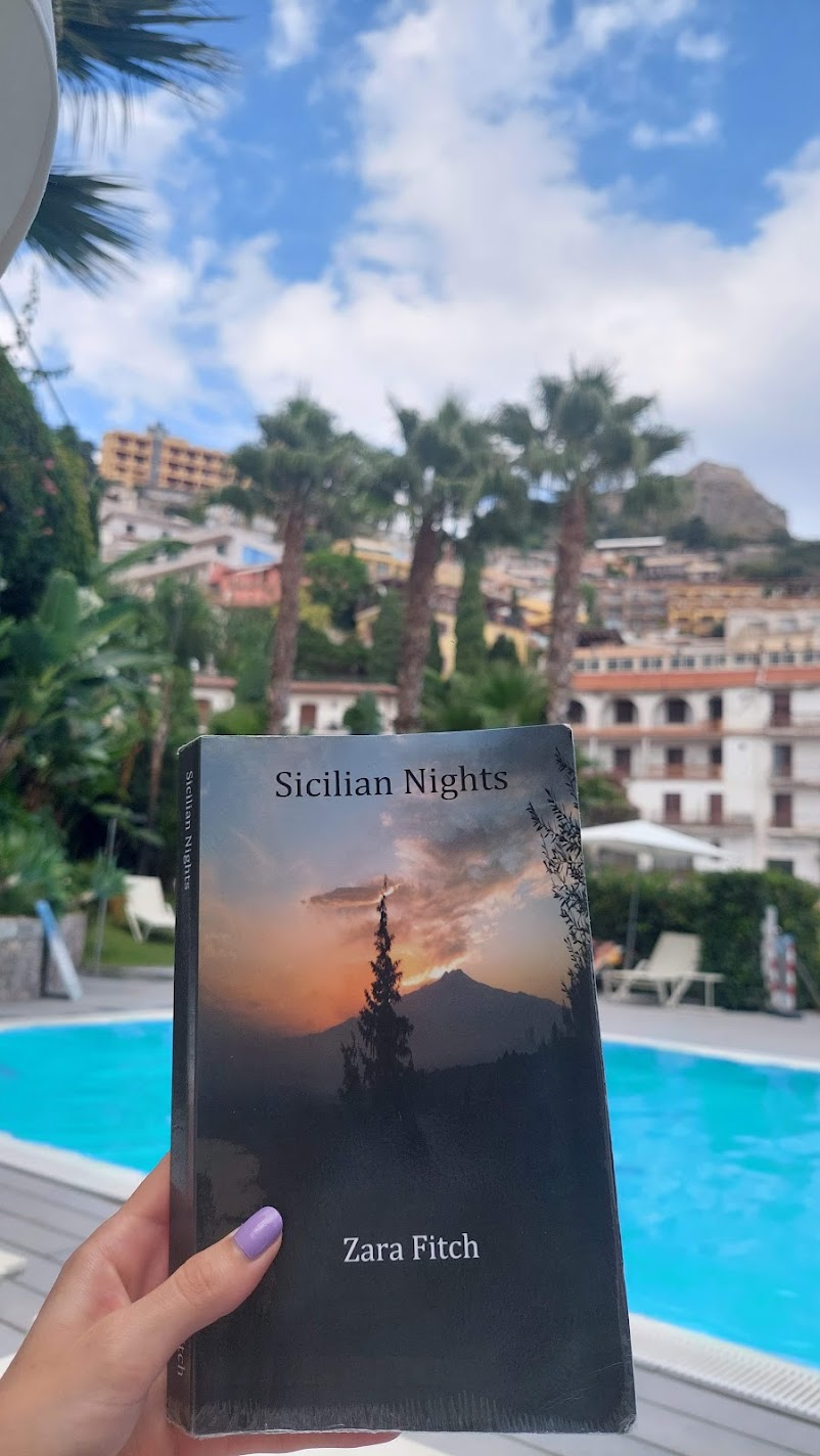 A hand holding the book "Sicilian Nights" by a hotel pool in Taormina
