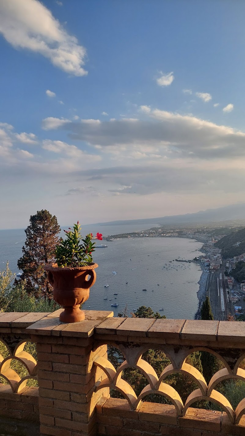 A terracotta urn with a red flower on a brick balustrade overlooking Taormina bay at golden hour