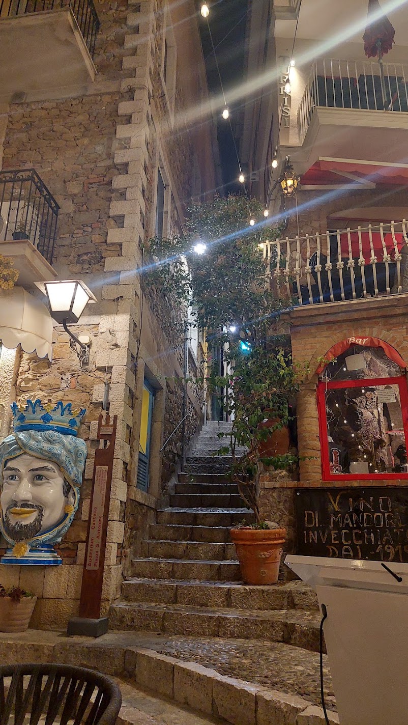 A stepped alley in Castelmola at night with a ceramic Moor's head and an almond-wine bar