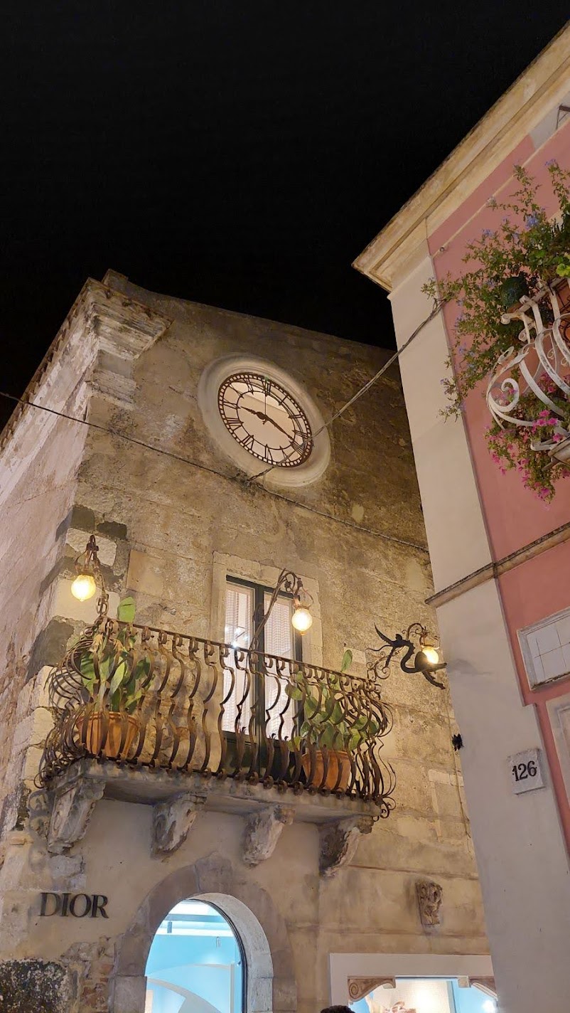A Baroque clock tower lit up at night above a boutique in Taormina, with a cactus-lined balcony