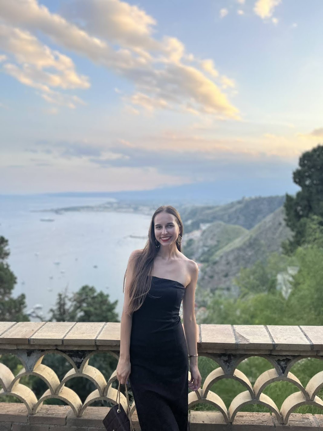 A woman in a black dress on the Villa Comunale terrace overlooking Taormina bay at dusk