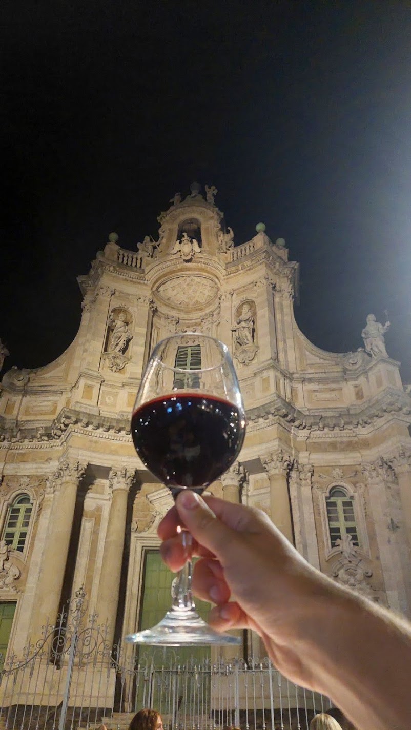 A hand holding a glass of red wine in front of the floodlit Basilica della Collegiata in Catania