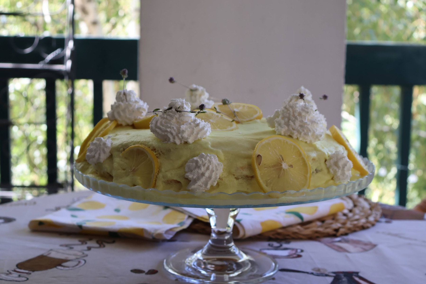 Whole lemon cake on a glass cake stand on a balcony table, with greenery blurred in the background
