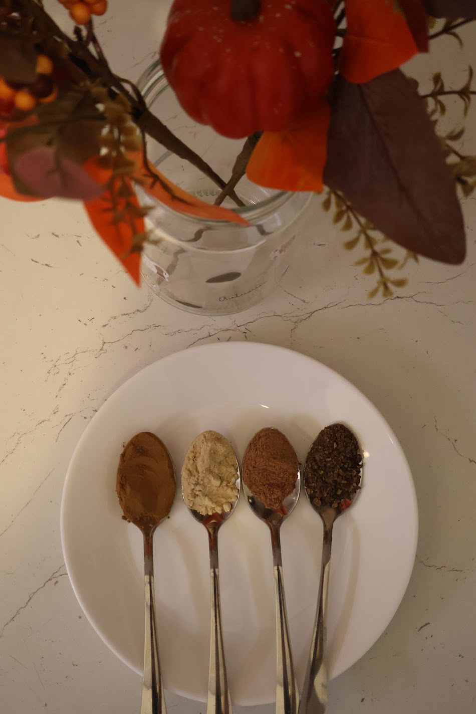 Four spoons of ground cinnamon, ginger, nutmeg and allspice on a white plate, with an autumn bouquet in a glass jar behind