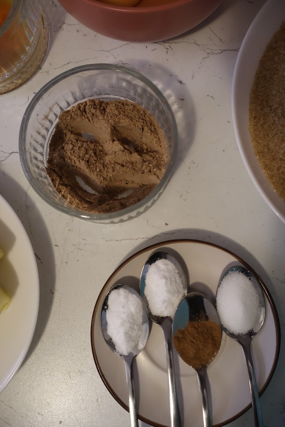 A glass bowl of homemade pumpkin spice beside a saucer of spoons, three with a white powder and one with a brown spice