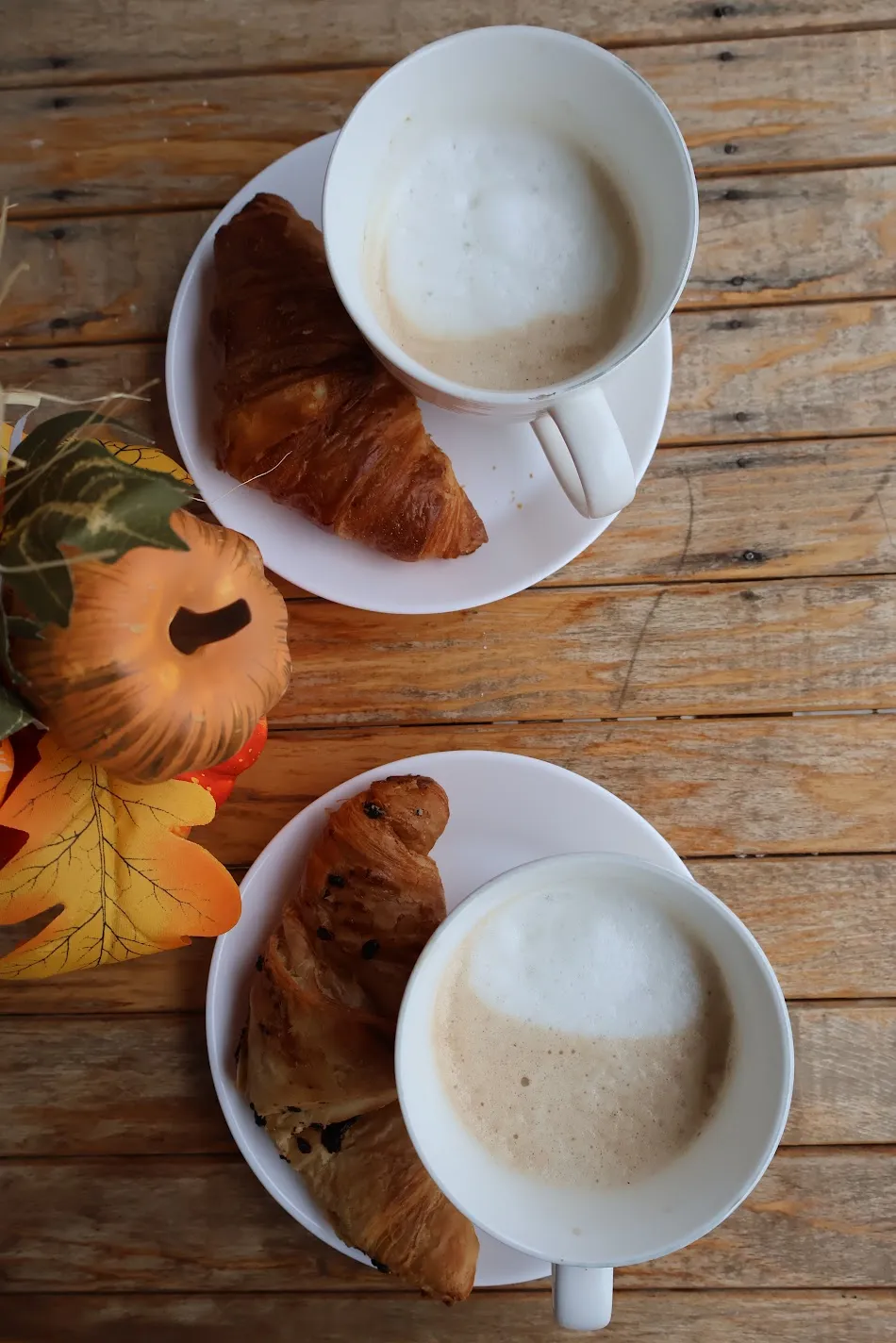 Two cappuccinos with croissants on a wooden table with autumn decorations, seen from above