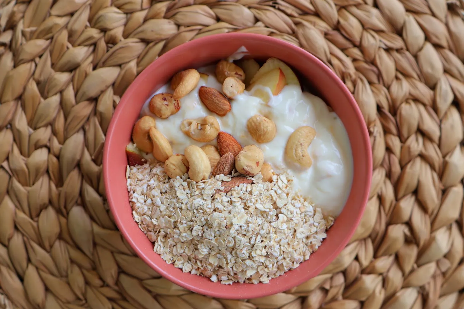Pink bowl of yogurt with oats, cashews, almonds and apple on a woven placemat