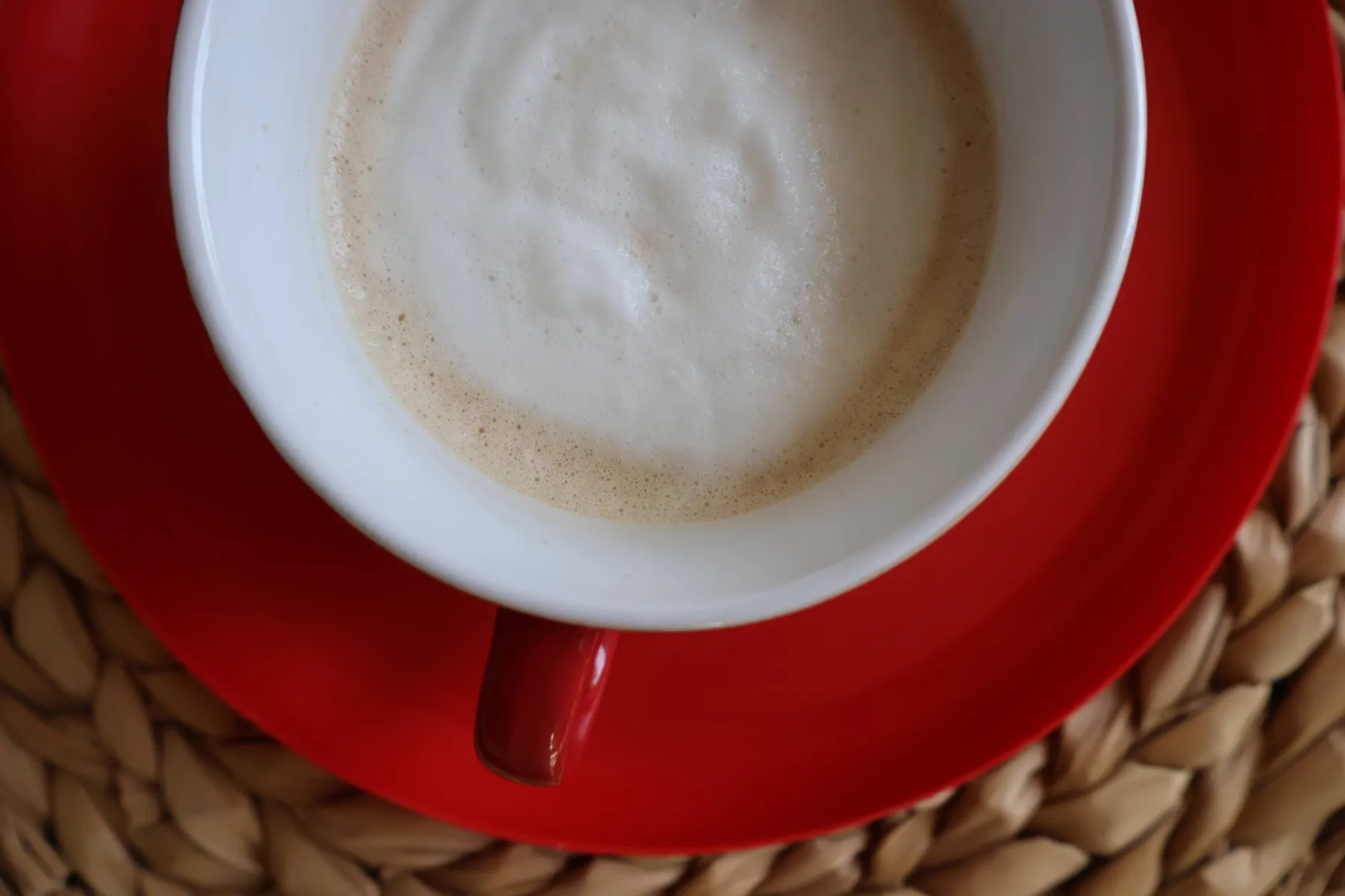 Cappuccino in a white cup on a red saucer, on a woven placemat, seen from above
