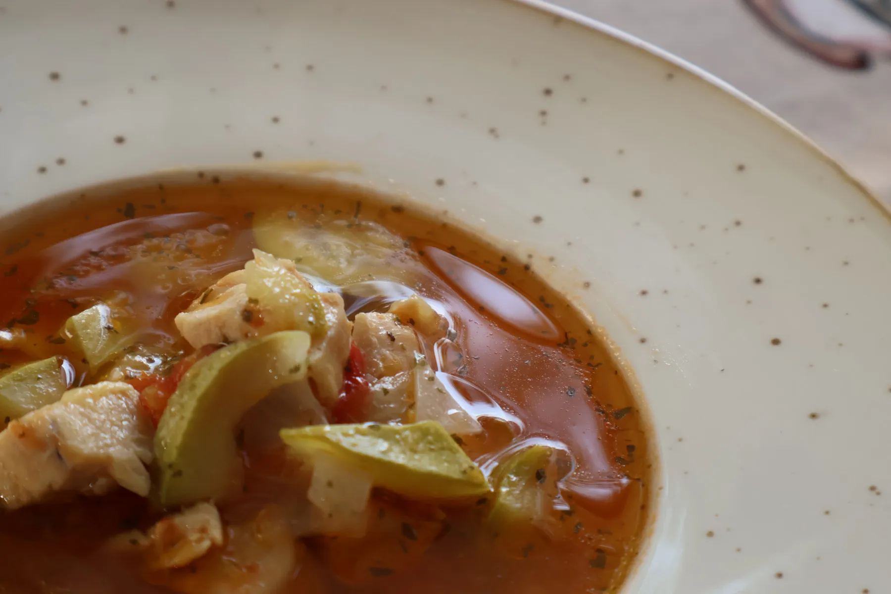 Close-up of chicken, courgette and tomato in a red broth with herbs, in a cream speckled plate