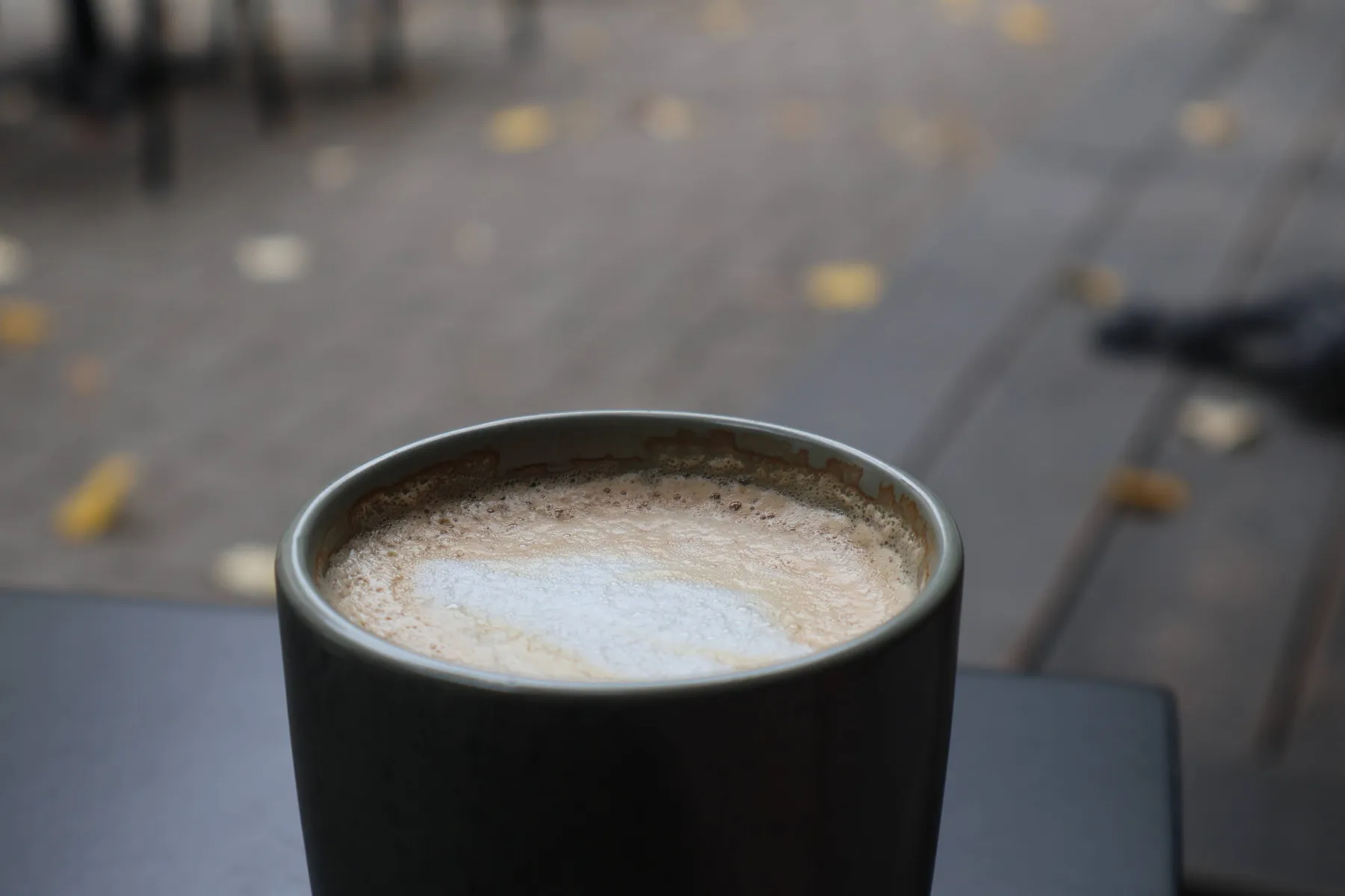 Macchiato in a dark grey cup on a café table, with fallen yellow leaves on the ground behind